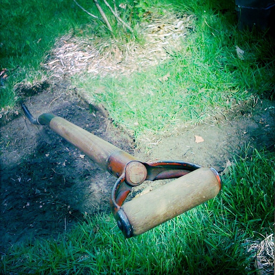 Hand-held sod cutting tool lying on grass next to a patch where turf has been lifted to bare soil