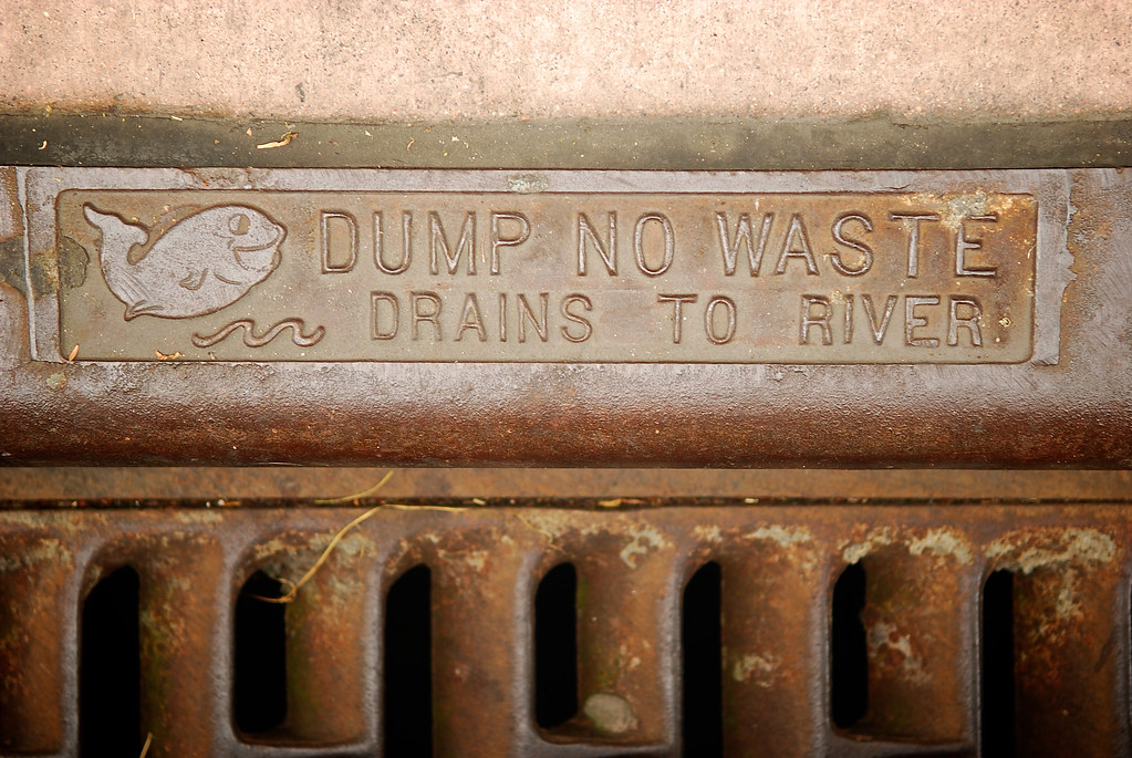 Rusty street storm drain grate cast with a fish symbol and the words Dump No Waste, Drains to River